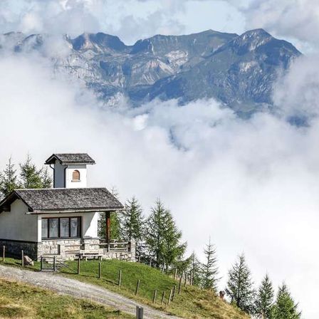 Ein kleines Kapellengebäude steht auf einem Hügel, umgeben von üppigen grünen Bäumen. Die Landschaft zeigt einen entfernten Gebirgszug mit nebligen Wolken.