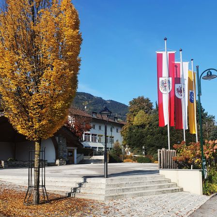 Ein Freibereich mit einem gelben Baum, Steinstufen und Fahnen an Stangen. Hinter dem Baum befindet sich ein Gebäude mit einer Treppe, die zum Eingang führt. Eine Straßenlaterne steht links vom Baum. In der Ferne sind Bäume und Berge zu sehen.