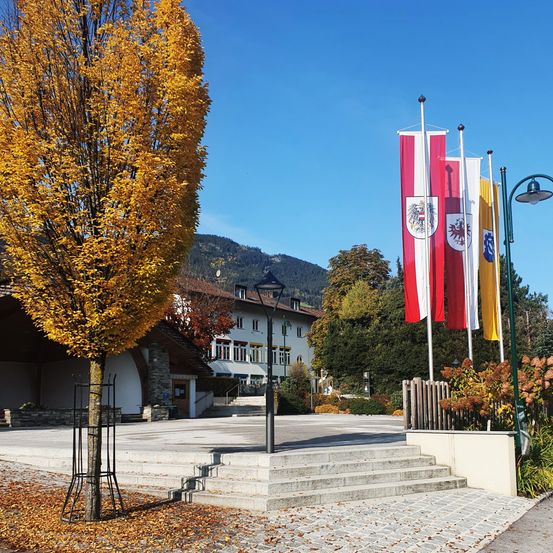 Ein Freibereich mit einem gelben Baum, Steinstufen und Fahnen an Stangen. Hinter dem Baum befindet sich ein Gebäude mit einer Treppe, die zum Eingang führt. Eine Straßenlaterne steht links vom Baum. In der Ferne sind Bäume und Berge zu sehen.