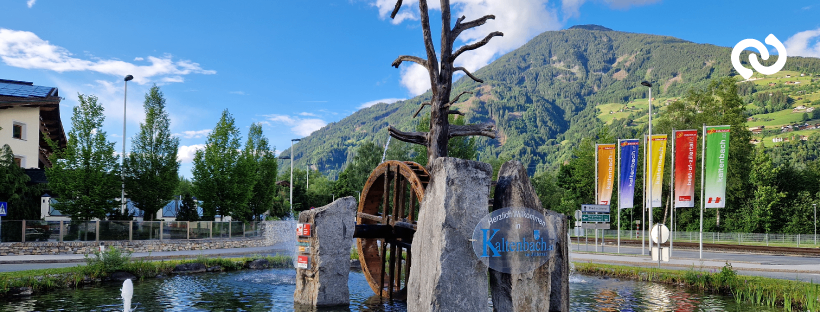 Ein szenischer Brunnen mit einem großen Holzrad in der Mitte, umgeben von Felsen und einem Baum, vor einer Kulisse von Bergen und einem klaren blauen Himmel. Bunte Banner sind am Straßenrand aufgestellt.