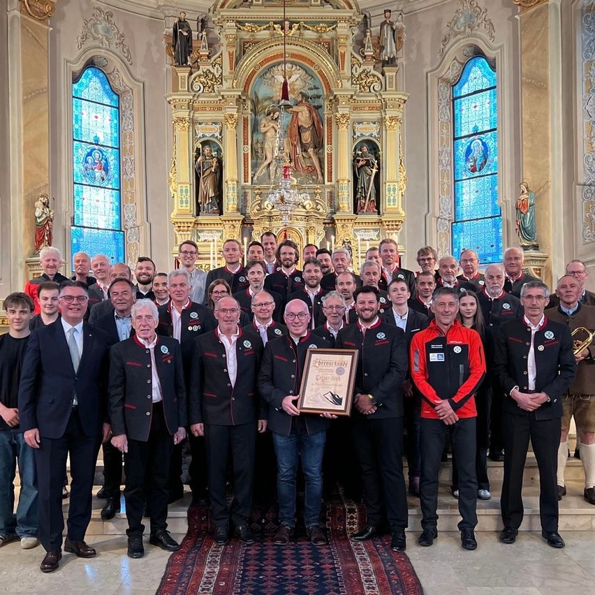 Eine Gruppe von Menschen in gleichen Uniformen posiert für ein Foto in einer Kirche. Ein Mann hält ein gerahmtes Zertifikat. Hinter ihnen ist ein dekorierter Altar mit Statuen zu sehen.