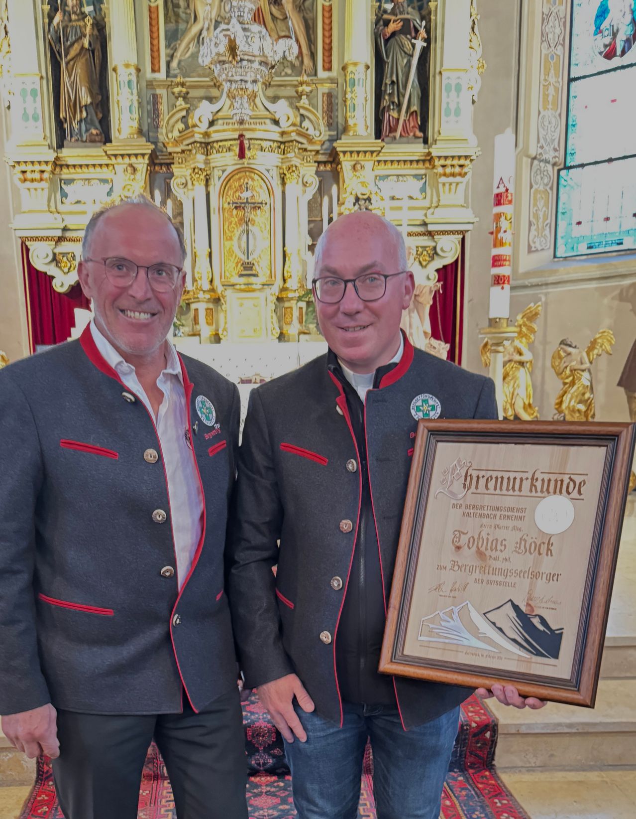 Zwei Männer in traditionellen Uniformen stehen vor einem Kirchenaltar, einer hält eine Plakette. Sie lächeln für die Kamera. Hinter ihnen schmücken ein goldener Altar und Statuen die Kirche.
