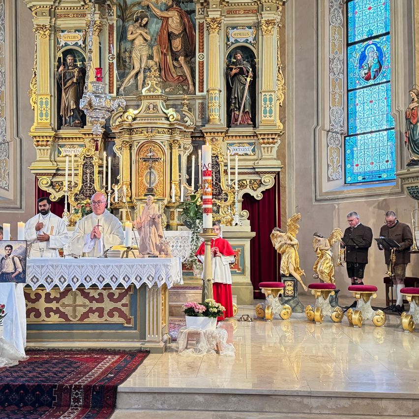Ein Priester zelebriert an einem Altar in einer dekorierten Kirche mit goldenen Statuen, Buntglasfenstern und Kerzen. Messdiener in roten Roben und andere sind anwesend.