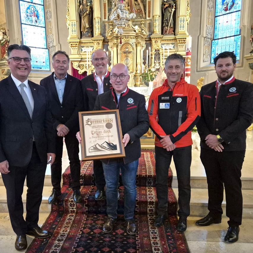 Fünf Männer stehen in einer Kirche und halten ein gerahmtes Zertifikat. Sie lächeln und scheinen für ein Foto zu posieren. Der Mann in der Mitte hält das Zertifikat. Der Mann auf der rechten Seite trägt eine orange Jacke, während die anderen schwarze Anzüge tragen.