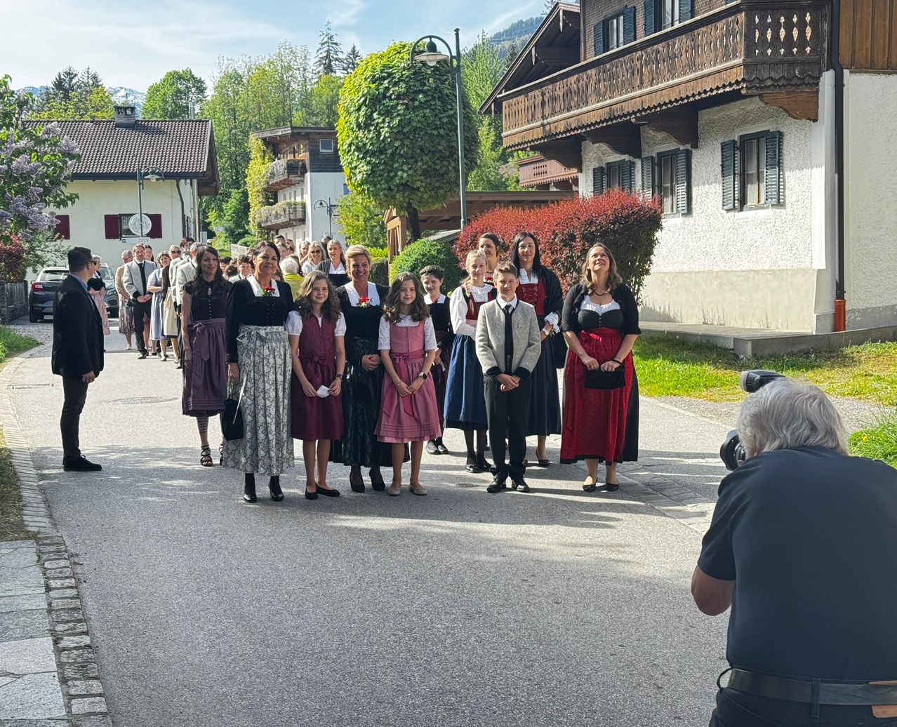 Eine Gruppe von Menschen in traditioneller Kleidung steht auf einer Dorfstraße. Ein Fotograf hält den Moment von der Seite fest. Im Hintergrund sind Gebäude und Grünflächen zu sehen.