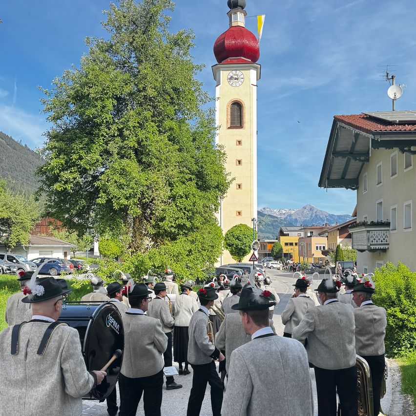 Eine Blaskapelle marschiert eine Straße entlang, gesäumt von Gebäuden und Bäumen. Im Hintergrund befindet sich ein Turm mit einer roten Kuppel.