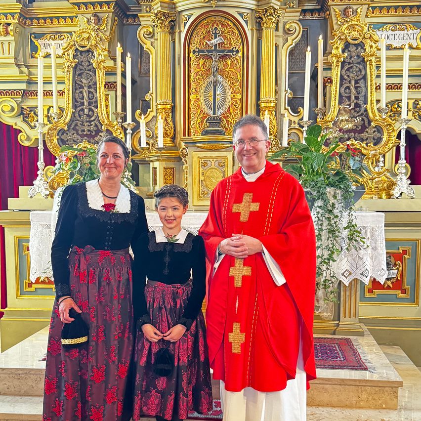 Ein Priester in Rot steht mit zwei Frauen in traditioneller Kleidung vor einem kunstvollen Altar.
