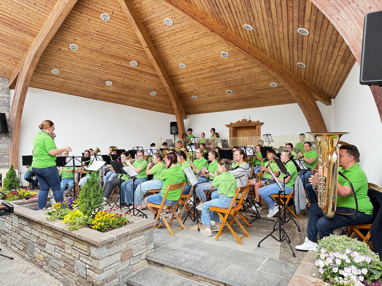 Ein Blechbläserensemble in grünen T-Shirts spielt in einem hölzernen Saal mit einem Tonnengewölbe. Die Musiker sitzen auf Stühlen, und ein Dirigent steht vor ihnen. Die Instrumente sind auf Notenständern platziert, und der Saal ist mit Pflanzen dekoriert.