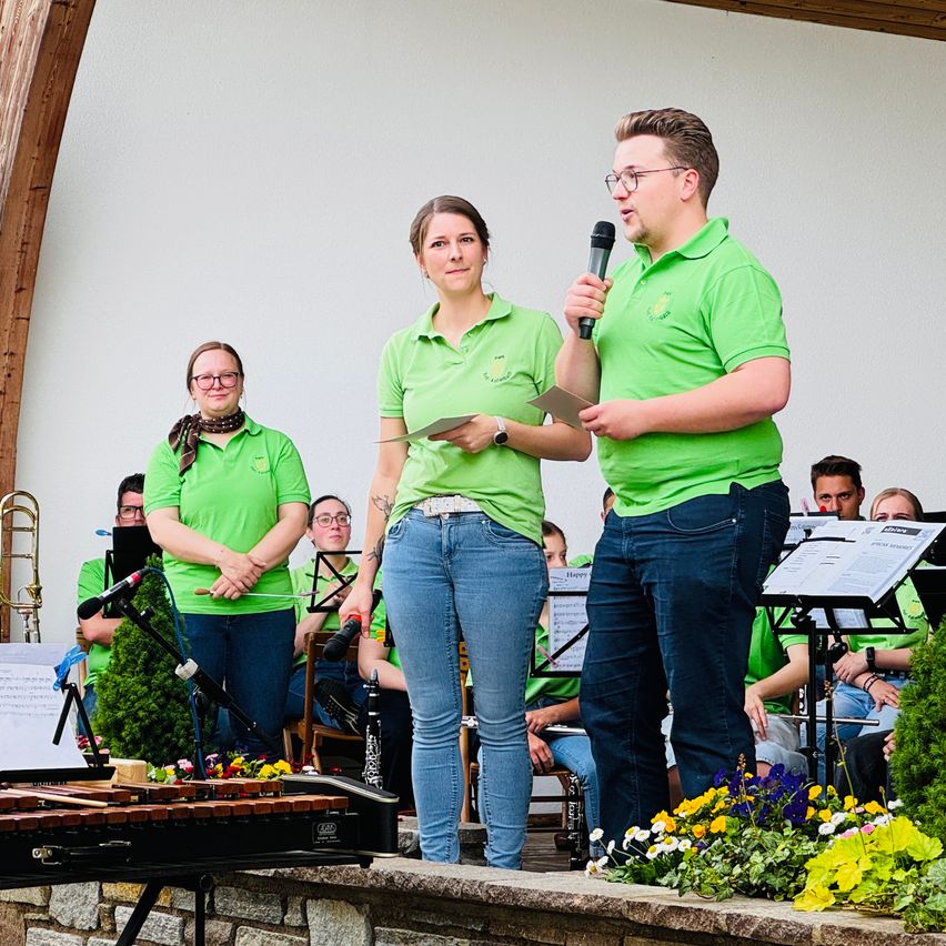 Zwei Personen stehen auf der Bühne, eine mit einem Mikrofon, und sprechen das Publikum an. Eine Frau vor ihnen hält ein Stück Papier in der Hand. Dahinter steht eine Band mit Instrumenten und Notenblättern.