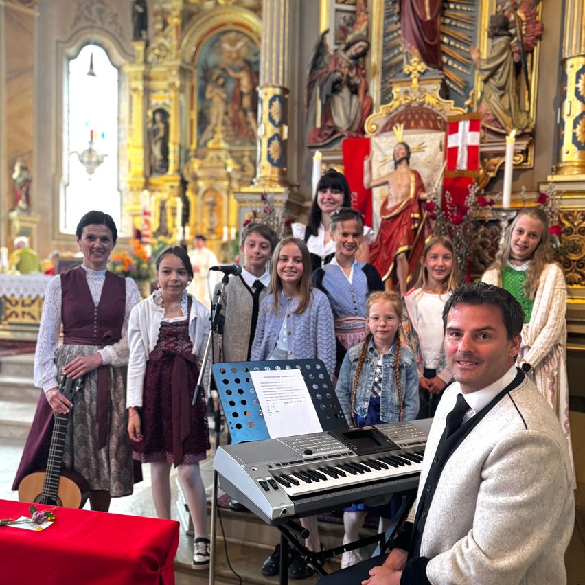 Ein Chor von Kindern in traditioneller Kleidung, begleitet von einem Mann in Anzug, steht vor einem Altar mit Musikinstrumenten und lächelt für ein Foto.