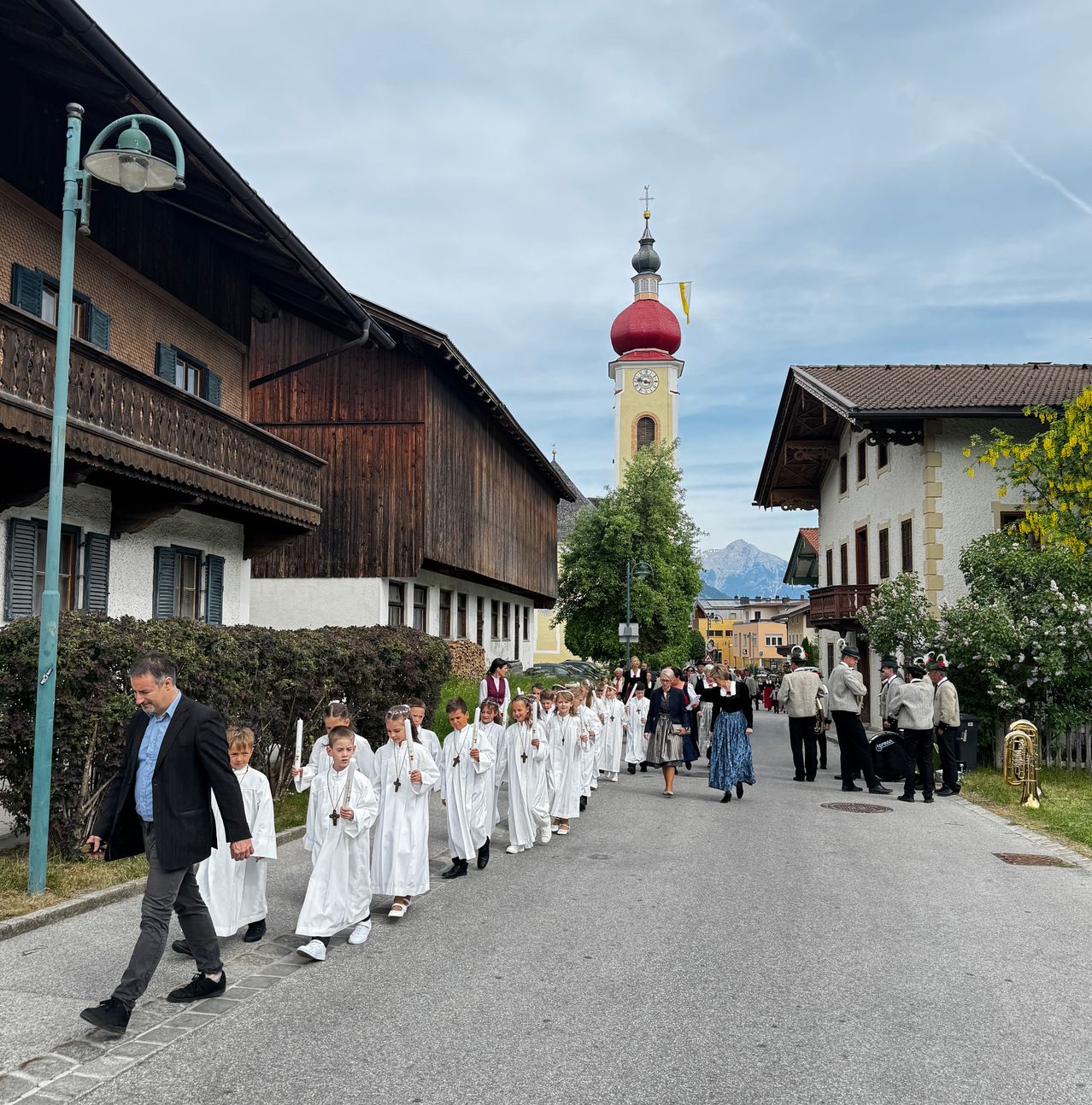 Eine Reihe von Kindern in weißen Gewändern geht eine Straße hinunter, gefolgt von einem Mann. Dahinter geht eine Gruppe von Menschen, einige in traditioneller Kleidung, in die entgegengesetzte Richtung. Im Hintergrund ist ein Kirchturm mit einer roten Kuppel zu sehen.