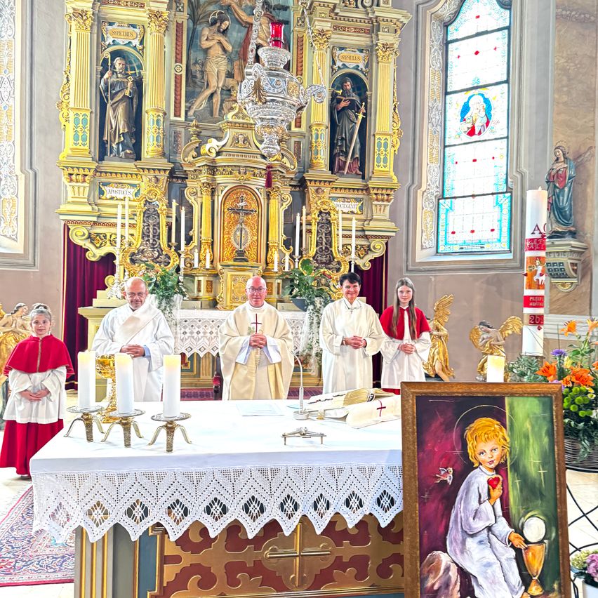 Priester stehen vor einem Altar in einer Kirche, geschmückt mit Kerzen und religiöser Kunst. Ein Gemälde eines Kindes, das einen Kelch und ein Herz hält, befindet sich in der Nähe.