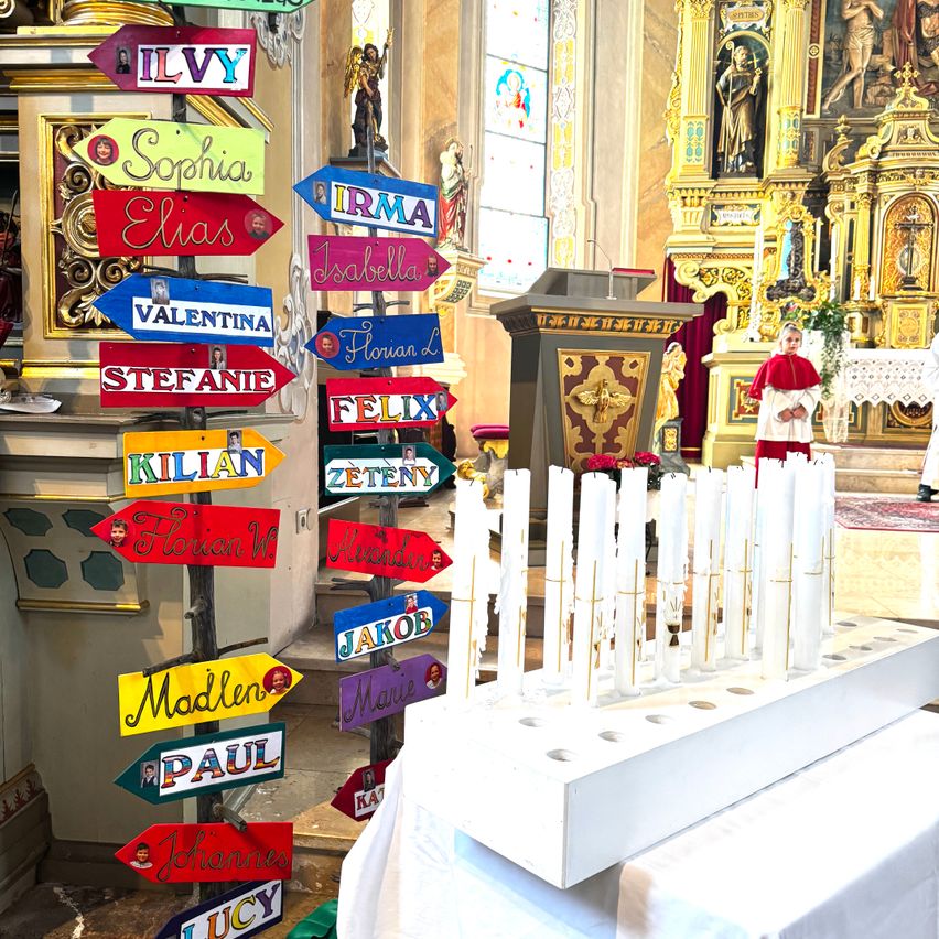 Der Innenraum einer Kirche zeigt farbenfrohe Schilder mit Kindernnamen und Fotos. Ein Junge in rotem Gewand steht neben einem Kerzenständer mit mehreren weißen Kerzen.