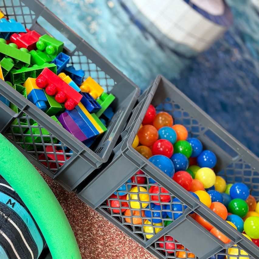 Zwei graue Plastikkisten voller Spielzeug. Eine ist mit bunten Bausteinen gefüllt, die andere mit bunten Bällen. In der Nähe liegt ein grüner Schwimmring.