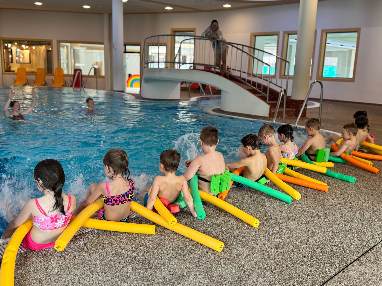 Mehrere Kinder sitzen auf bunten Schwimmhilfen in einem Pool, während eine Frau oben auf einer Treppe steht und zusieht.
