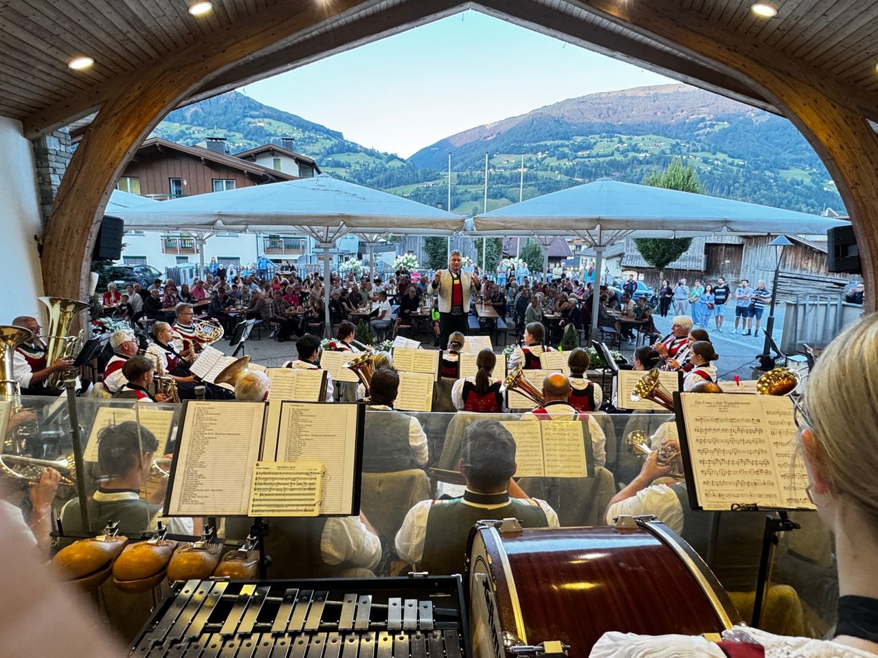Ein Blasorchester spielt auf der Bühne unter einem großen Schirm, während eine Menge vor einem malerischen Bergpanorama zuschaut.