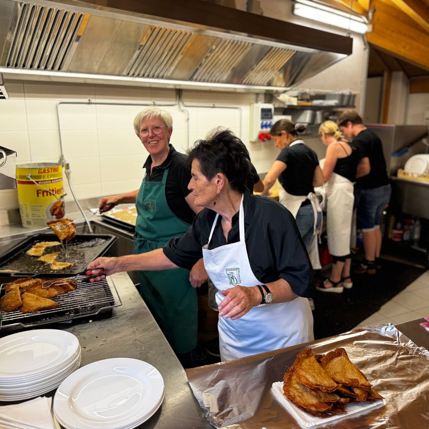 Eine Gruppe von Menschen in einer Küche, die Schürzen tragen, bereitet Essen zu. Eine Frau brät Essen auf einem Grill.