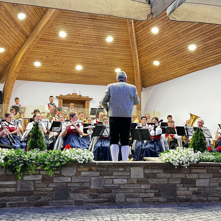 Ein Orchester spielt auf einer Bühne mit Holzdecke und Ziegelmauer, geschmückt mit weißen Blumen und Grün. Der Dirigent steht vorne, und Musiker spielen verschiedene Instrumente.