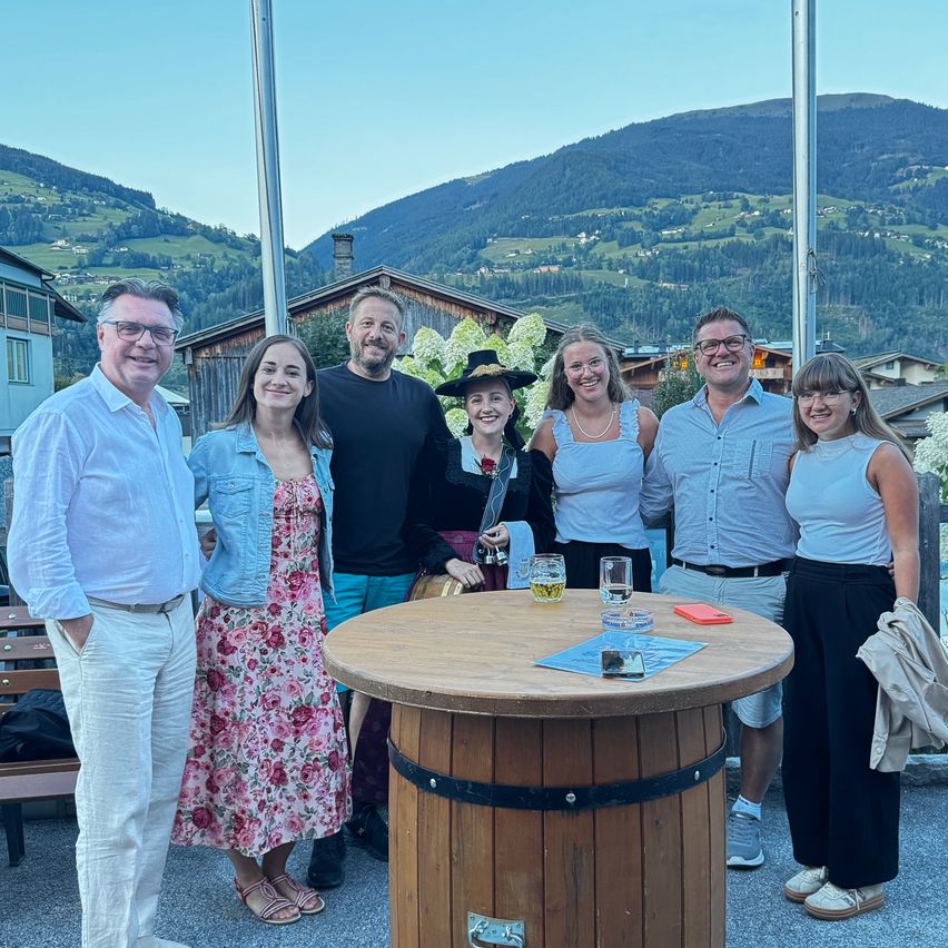 Eine Gruppe von Menschen steht um einen Holzfass herum, lächelt und posiert für ein Foto mit einem Berg im Hintergrund.