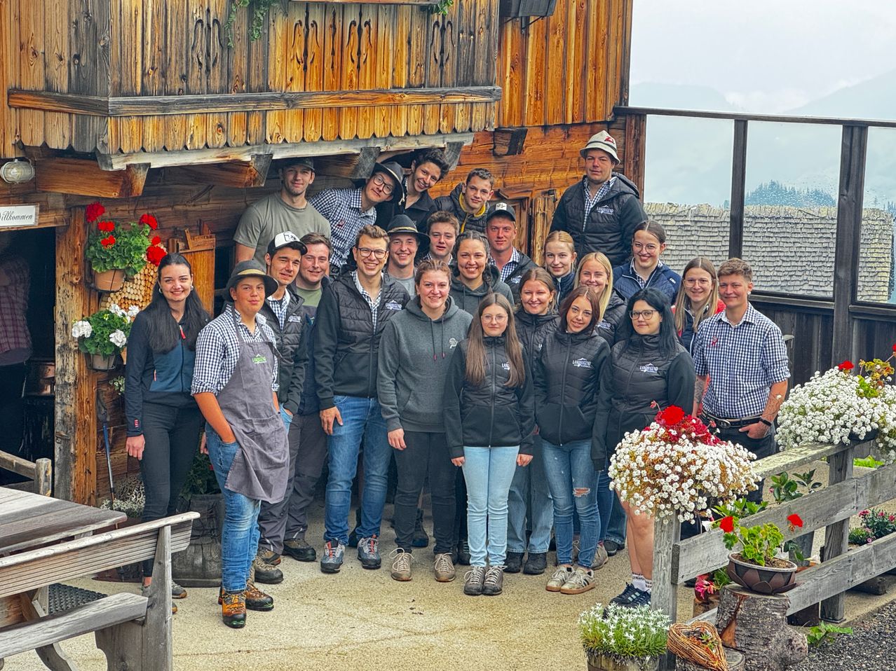 Eine Gruppe junger Menschen in Winterkleidung steht vor einem Holzhaus mit Blumen und Grünpflanzen um sie herum.