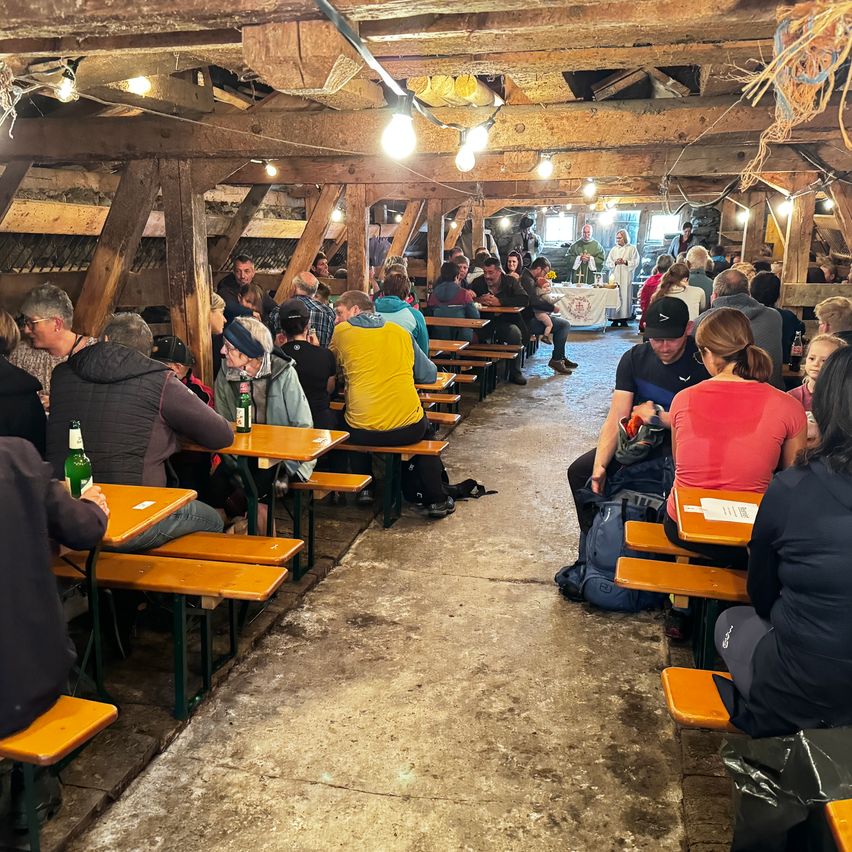 Eine Gruppe von Menschen sitzt an Tischen in einem rustikalen Holzspeisebereich mit Holzbalken und hängenden Lichtern. Einige tragen Hüte und Brillen, und einige haben Flaschen auf ihren Tischen. Eine Person in einem weißen Gewand steht in der Nähe eines Tisches mit einem weißen Tuch.
