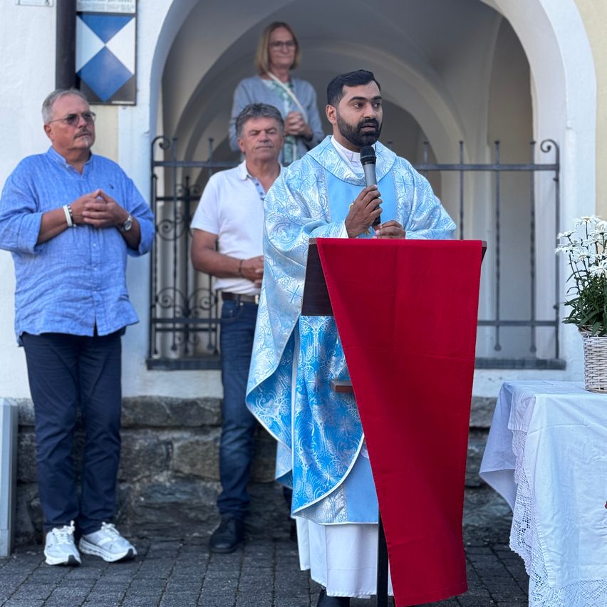 Ein Priester spricht in ein Mikrofon bei einer Outdoor-Veranstaltung, mit zwei Männern hinter ihm und einer Frau im Hintergrund.