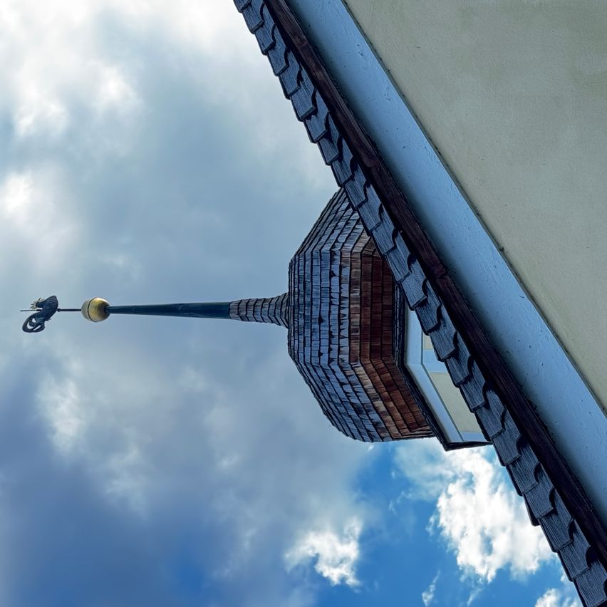 Ein Nahaufnahme eines Daches mit einem Kirchturm, einer Wetterfahne und Schindeln unter einem bewölkten Himmel.