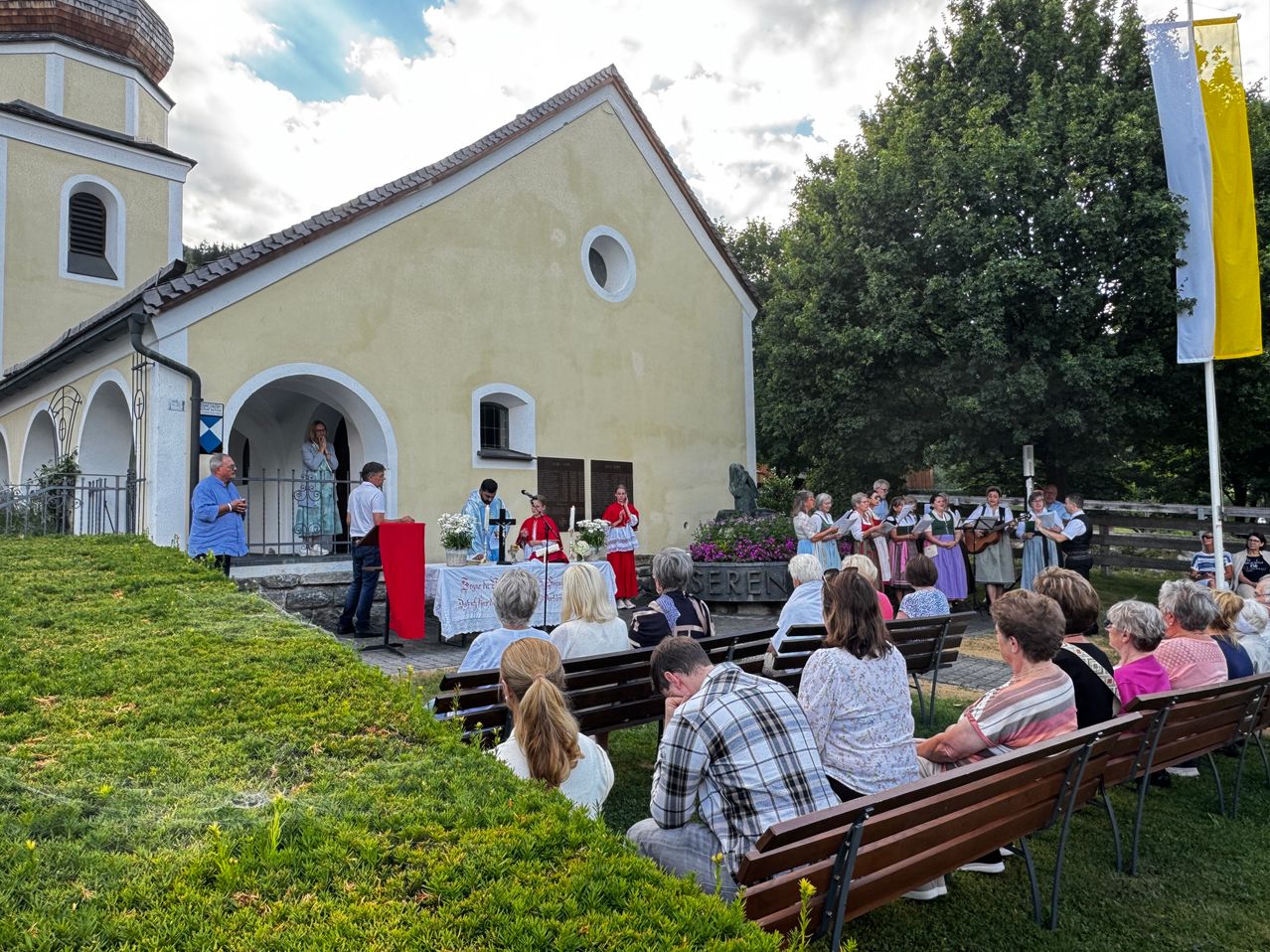 Eine Gruppe von Menschen versammelt sich vor einer Kirche für eine Zeremonie. Einige Personen stehen in der Nähe des Altars, während andere auf Bänken sitzen. Ein Chor singt, während einige Personen Musikinstrumente halten.