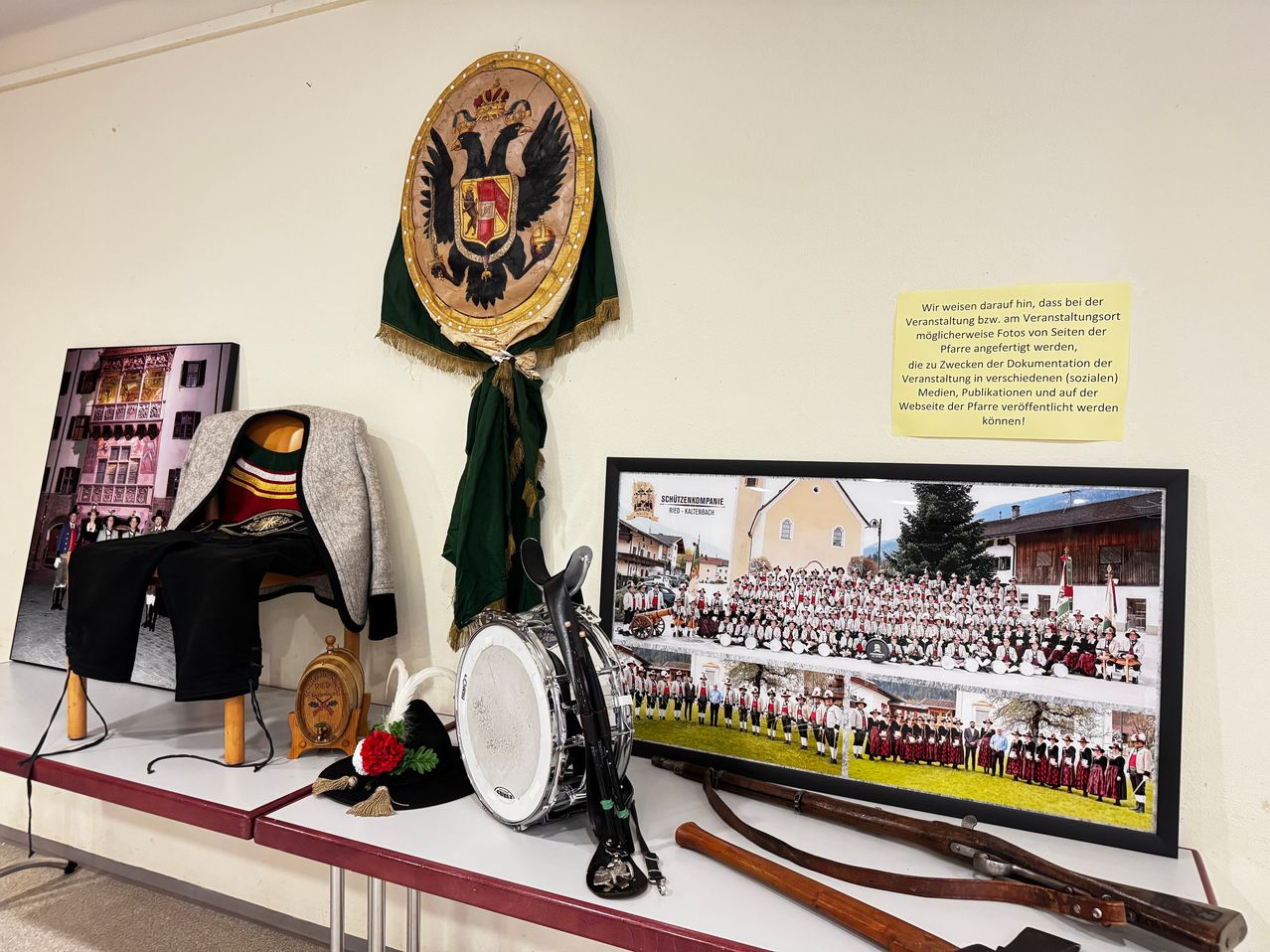Eine Ausstellung von traditionellen Kostümen und Musikinstrumenten auf einem Tisch. Ein gerahmtes Bild zeigt eine Gruppe von Menschen in Uniform, wahrscheinlich eine Blaskapelle. Ein Mantel und Hut mit einer Blume schmücken den Tisch. Ein dekoratives Emblem hängt an der Wand.