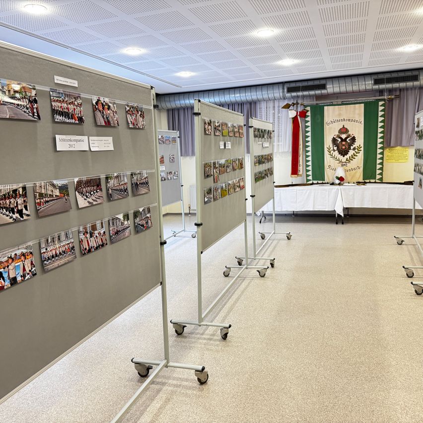 Ein Raum mit einer grauen Wand, die Fotos aus dem Jahr 2012 zeigt. Es gibt rollende Ständer mit Fotos und eine grüne Fahne mit Text. Ein mit einem weißen Tuch bedeckter Tisch steht im Raum.