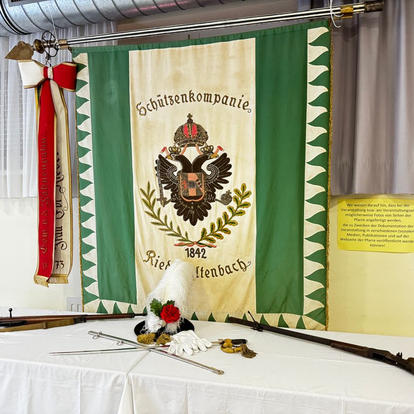 Eine grün-weiße Fahne mit einem Adlerwappen und Text hängt an einer Wand. Unter der Fahne sind auf einem Tisch historische Gegenstände wie Schwerter, eine Mütze und ein Gewehr ausgestellt.