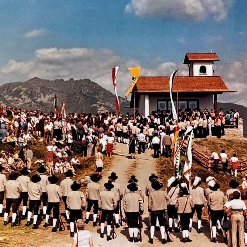 Eine Gruppe von Menschen in traditionellen Uniformen, die an einem sonnigen Tag mit Bergen im Hintergrund an einem Umzug teilnehmen, während eine Menge zuschaut.