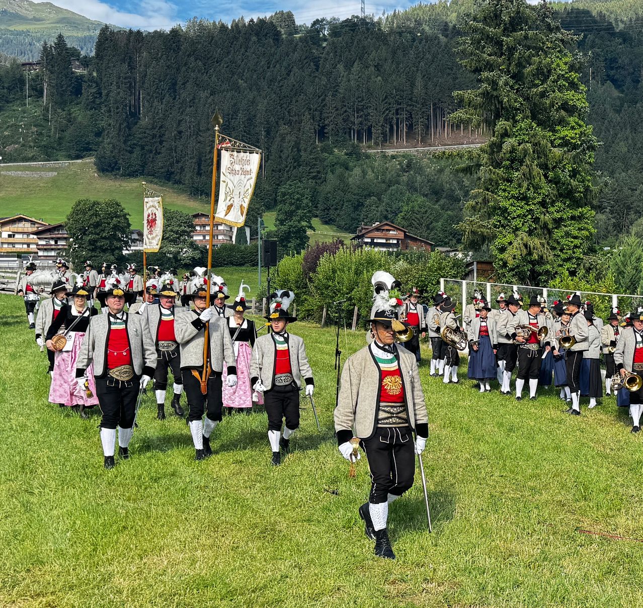 Eine Blaskapelle in traditioneller Kleidung tritt auf einem Freiluftfeld auf, umgeben von dichtem Wald und Bergen. Einige Mitglieder halten Banner und Flaggen.