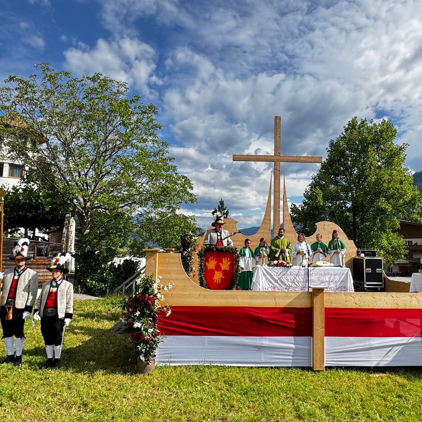 Eine Gruppe von Menschen in traditioneller grüner und weißer Kleidung versammelt sich um einen hölzernen Altar mit einem Kreuz in einem Berggebiet. Zwei Männer stehen als Wachen an der Seite des Altars.