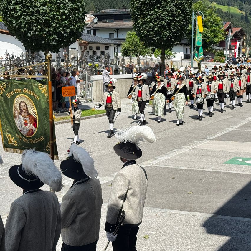 Ein Umzug von Menschen in traditionellen Kostümen mit Hüten und weißen Federn zieht die Straße hinunter, begleitet von Trommlern. Ein grünes Banner mit einem Bild von Jesus befindet sich links.