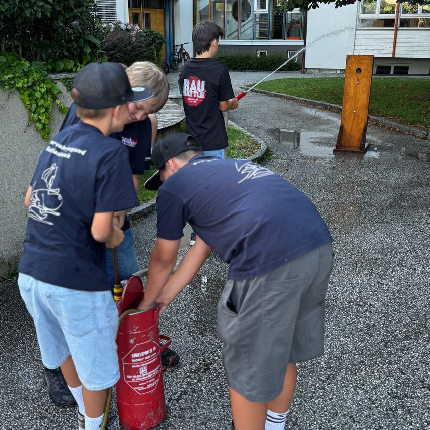 Drei Jungen füllen einen Feuerlöscher mit Wasser, einer hält den Schlauch, ein anderer hält den Feuerlöscher und der dritte beobachtet. Sie stehen vor einem Gebäude, in dessen Nähe ein Fahrrad geparkt ist.