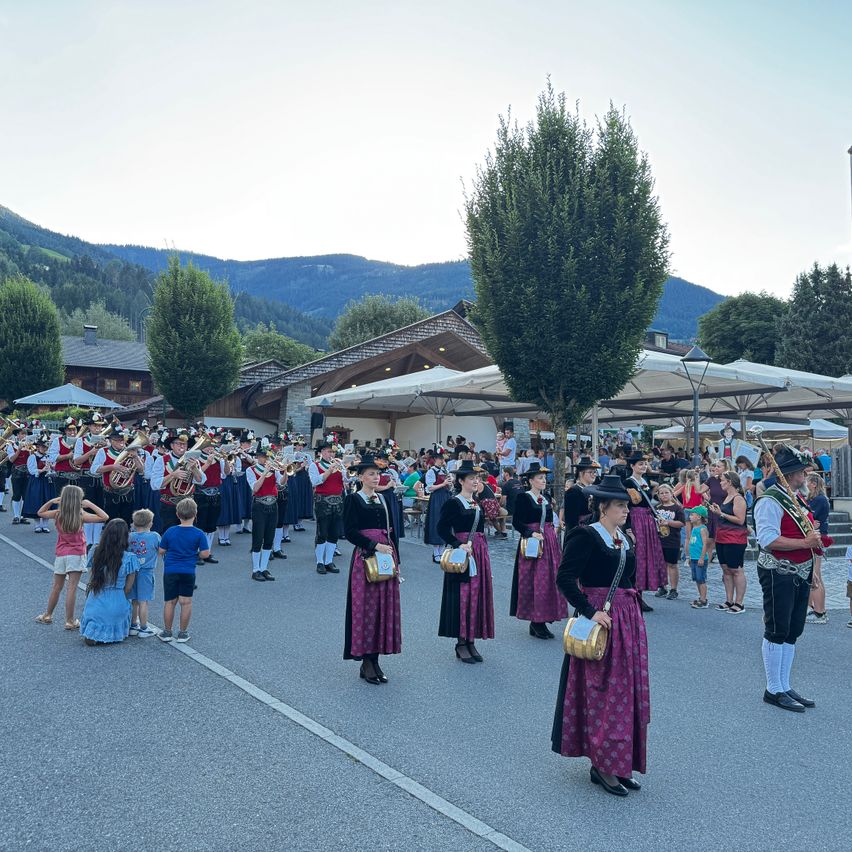 Eine Blaskapelle in traditioneller Kleidung tritt auf einer Straße auf, während Zuschauer und Kinder zuschauen. Im Hintergrund befinden sich Berge.
