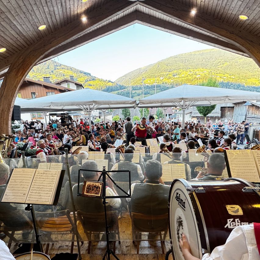 Eine große Menschenmenge versammelt sich unter einem Baldachin für eine musikalische Aufführung, mit Musikern, die verschiedene Instrumente spielen, und einer malerischen Bergkulisse.