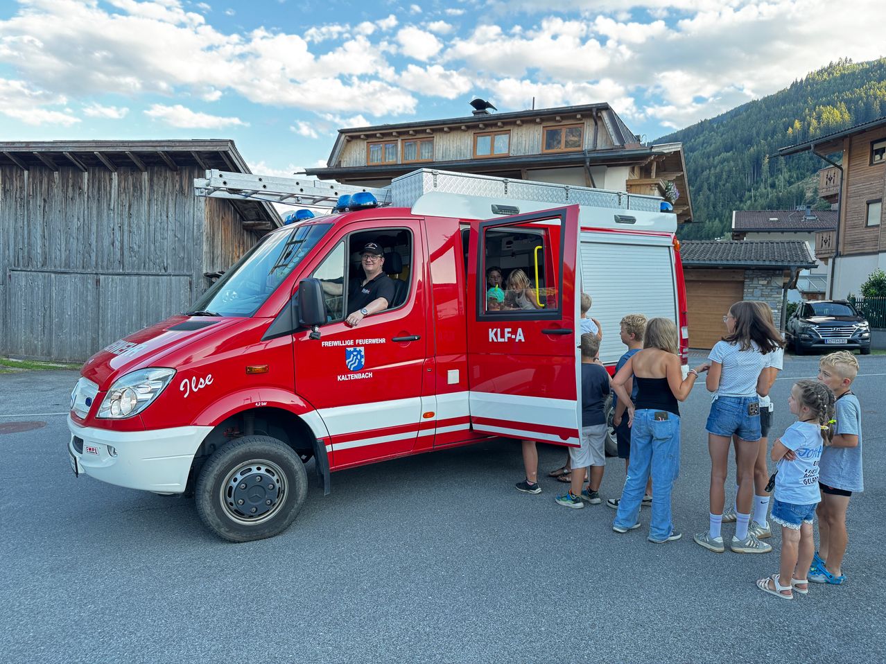 Ein roter Feuerwehrwagen mit der Aufschrift 'KLF-A' steht vor einem Gebäude, in das mehrere Kinder sehen.