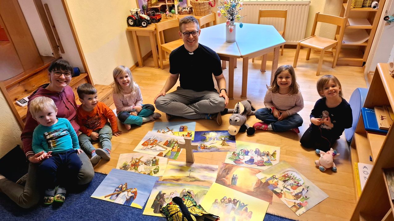 Ein Mann und drei Kinder sitzen auf dem Boden mit Illustrationen biblischer Geschichten. Der Mann trägt eine Brille und ein schwarzes T-Shirt. Spielzeug und ein kleines Kreuz liegen auf dem Boden.