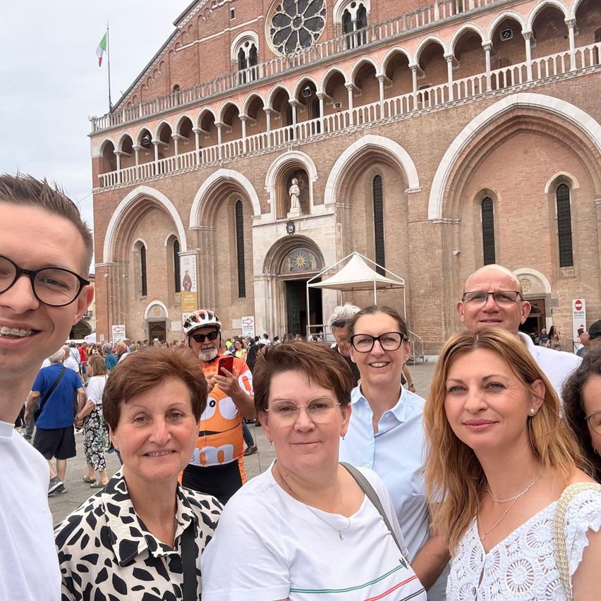 Eine Gruppe von Menschen posiert für ein Foto vor einem großen, prächtigen Gebäude. Sie lächeln und das Gebäude hat Bogenfenster und einen Glockenturm.