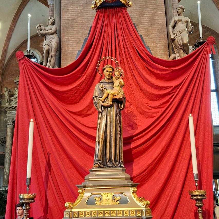 Eine Statue einer Frau, die ein Kind hält, steht auf einem Podest mit goldenen Verzierungen. Dahinter verdecken zwei große rote Vorhänge eine Ziegelmauer. Auf beiden Seiten der Statue befinden sich brennende Kerzen in goldenen Haltern. Über den Vorhängen stehen zwei Statuen von Frauen, die ein Baby halten.