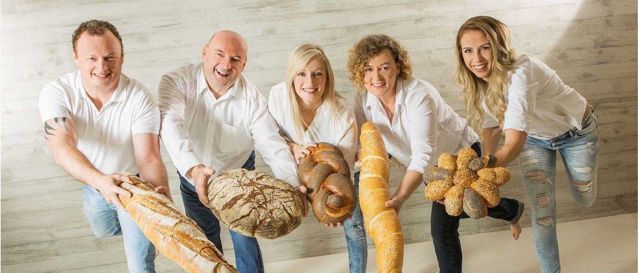 Fünf Menschen posieren mit Brot in einem Raum und lächeln. Der Mann auf der linken Seite hält ein Brot. Ein Logo für Adolf Wild ist unten links sichtbar.