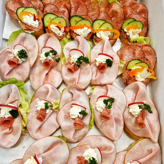 Eine Schachtel gefüllt mit verschiedenen Sandwiches aus dünnen Schinken- und Salamischeiben, garniert mit Frischkäse, Gurken und anderen Gemüsesorten.