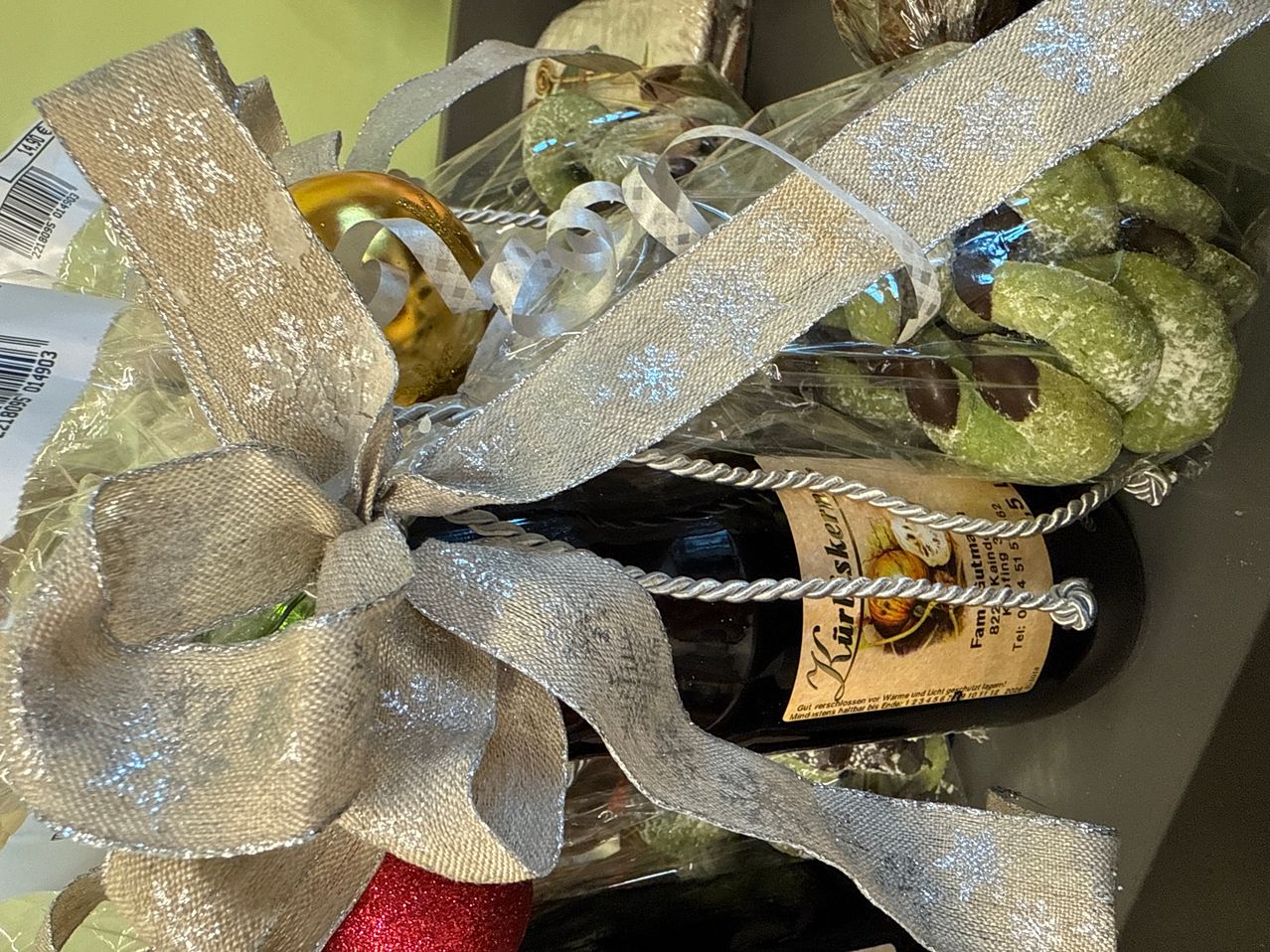 Eine Weinflasche, die in ein silbernes Band gewickelt ist, in einem Geschenkkorb mit einer goldenen Kugel, grünen Keksen und einem Juteseil platziert.
