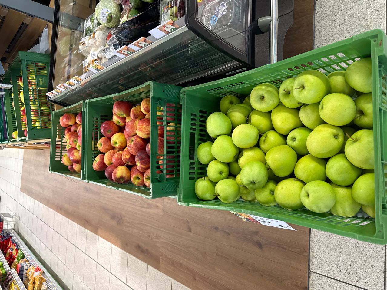 A supermarket aisle displays several green crates filled with apples and red crates with red apples, with a refrigerator in the background.