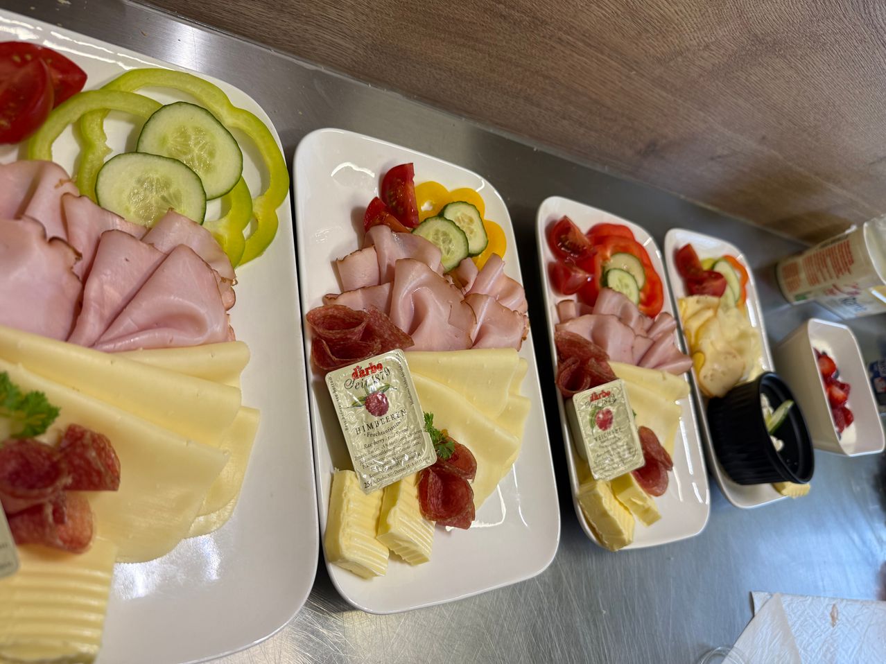 Vier weiße Teller mit einer Auswahl an geschnittenem Fleisch, Käse und Gemüse auf einem Holztisch. Jeder Teller enthält Schinken, Käse, Gurken und Tomaten.