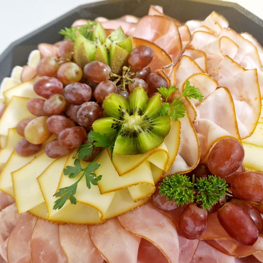 Ein Tablett zeigt eine Auswahl an Aufschnitten, Käse, Trauben und Kiwi. Die Aufschnitte sind geschnitten und angeordnet, begleitet von Käsescheiben. Trauben und Kiwi sind kunstvoll platziert.