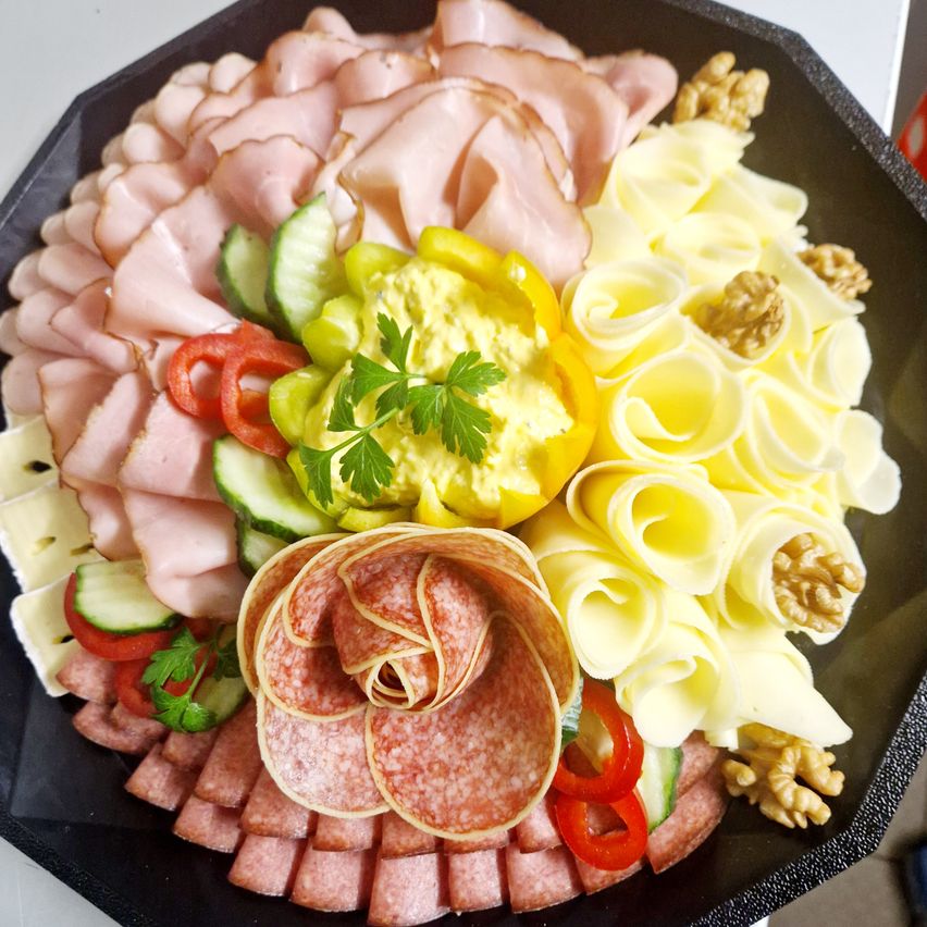 Ein großer Teller mit verschiedenen Fleischsorten, Käse, Gemüse und Nüssen. Schinkenscheiben, Salami, Käseräder, Gurken, Paprika und Walnüsse sind kunstvoll angeordnet.