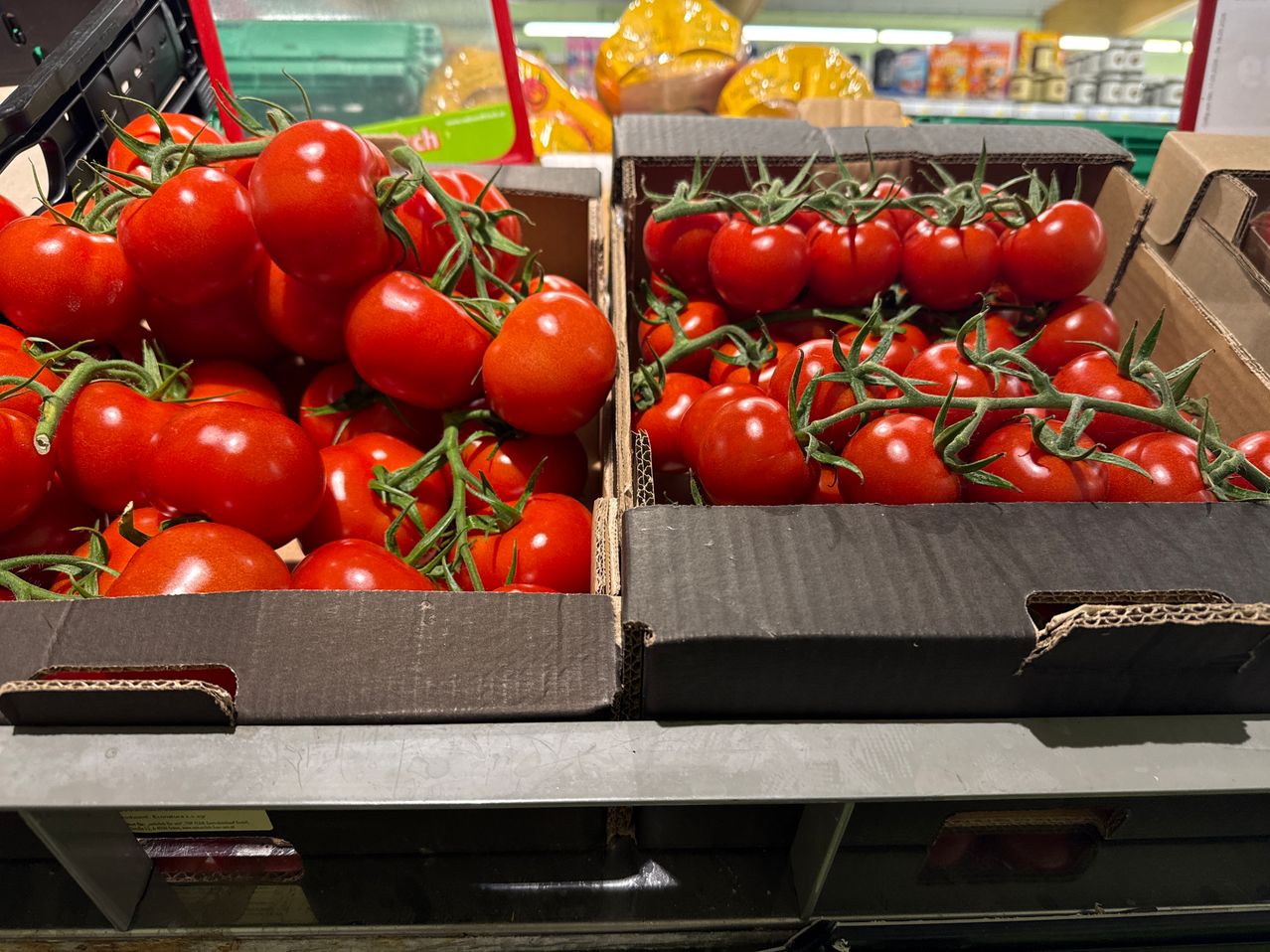 Two cardboard boxes filled with ripe red tomatoes on display in a supermarket, with shelves in the background.